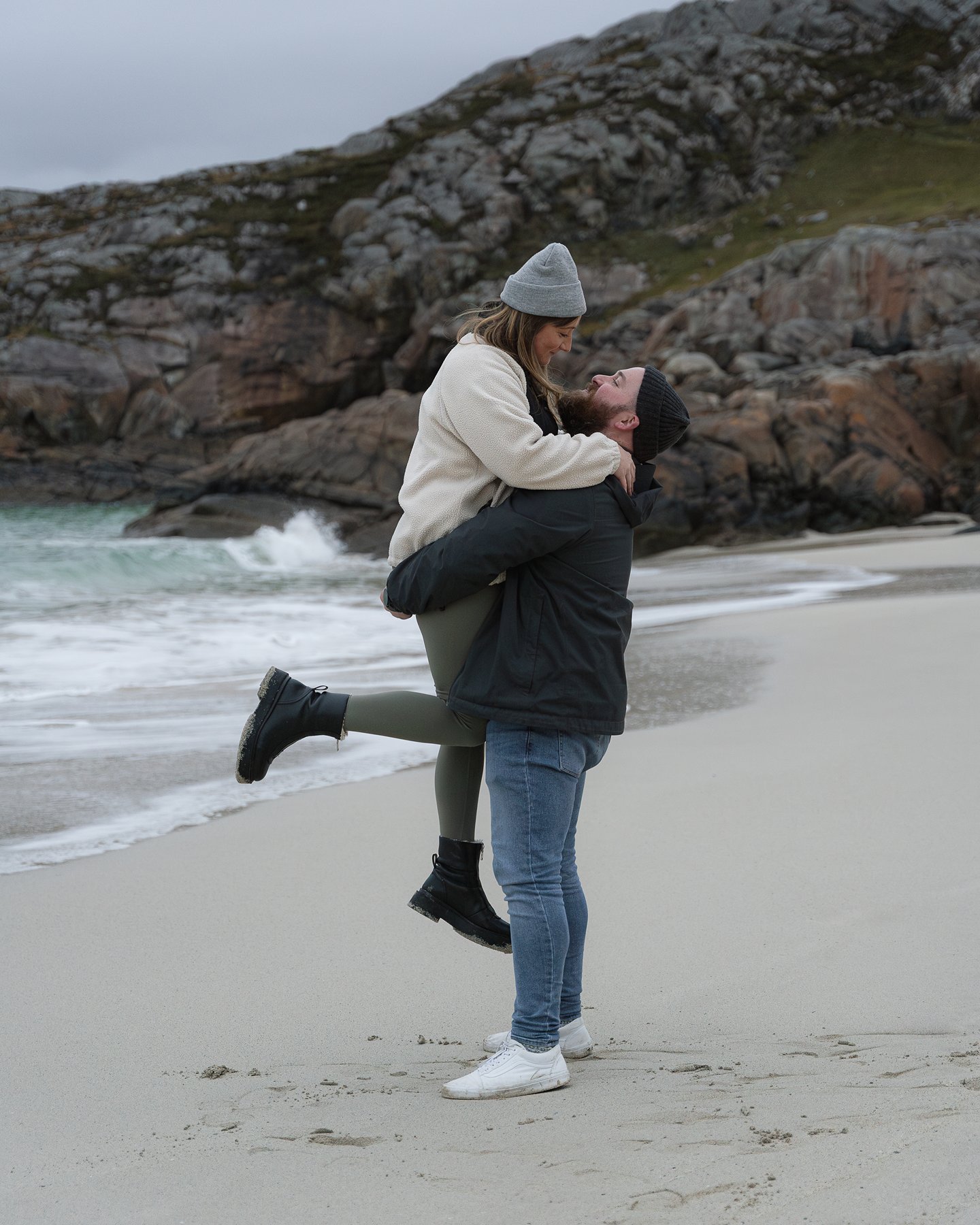 Man lifting a woman in his arms on a sandy beach, both smiling as waves crash nearby with rocky cliffs behind.