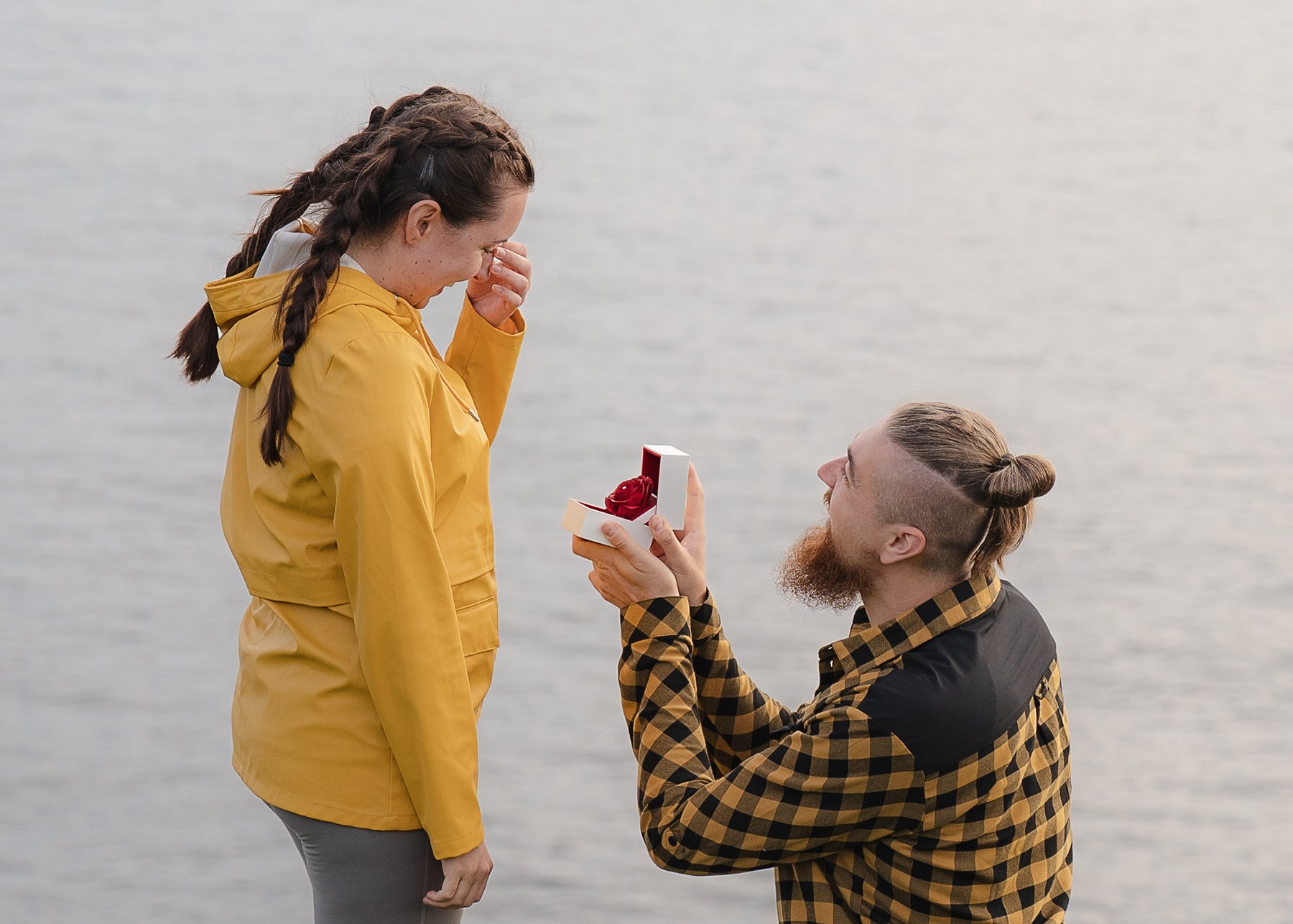 Man proposes on one knee by the water, holding an engagement ring box, as woman reacts emotionally in yellow jacket.