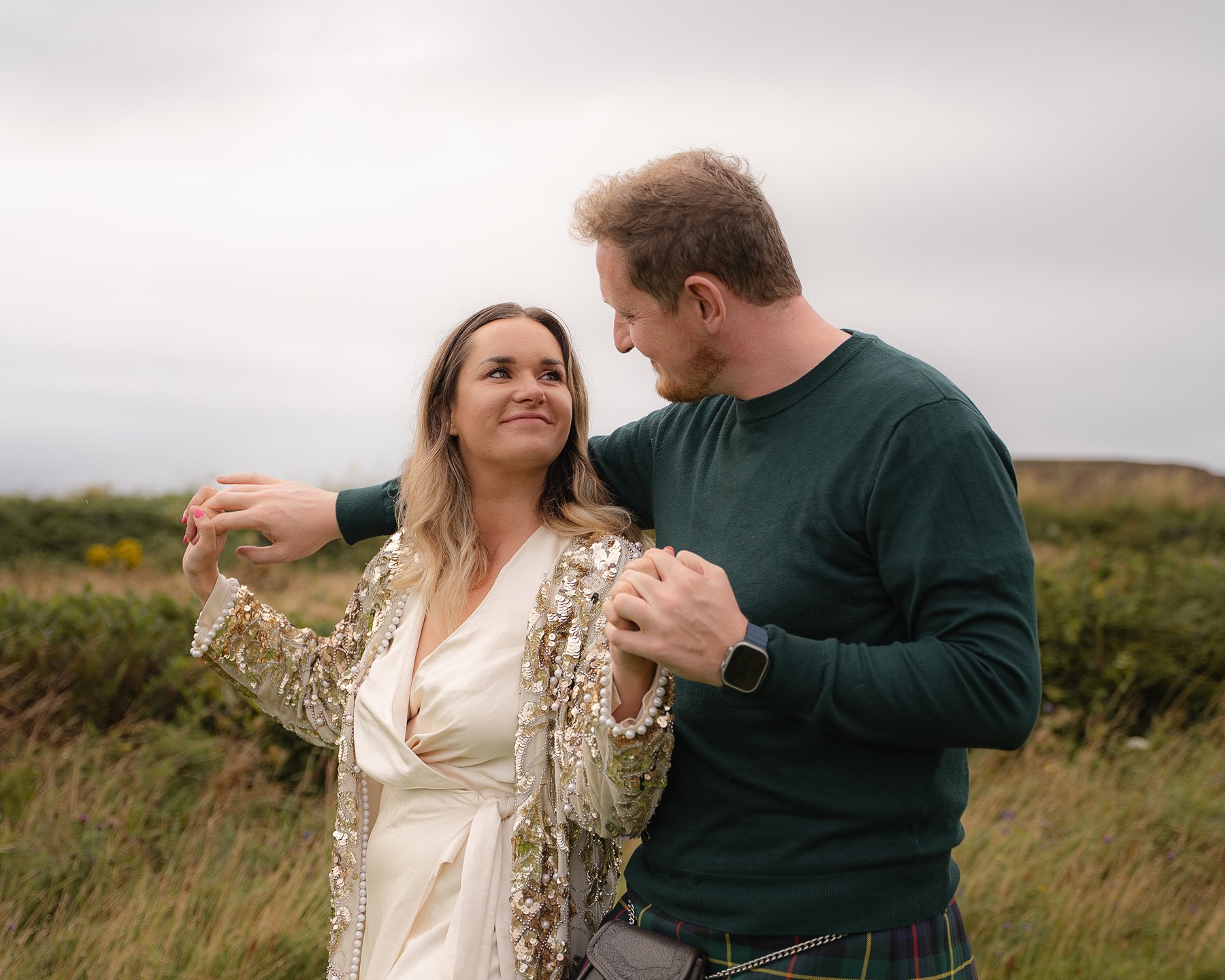 Couple holding hands and smiling at each other outdoors, woman in sequined jacket and man in green sweater with kilt.