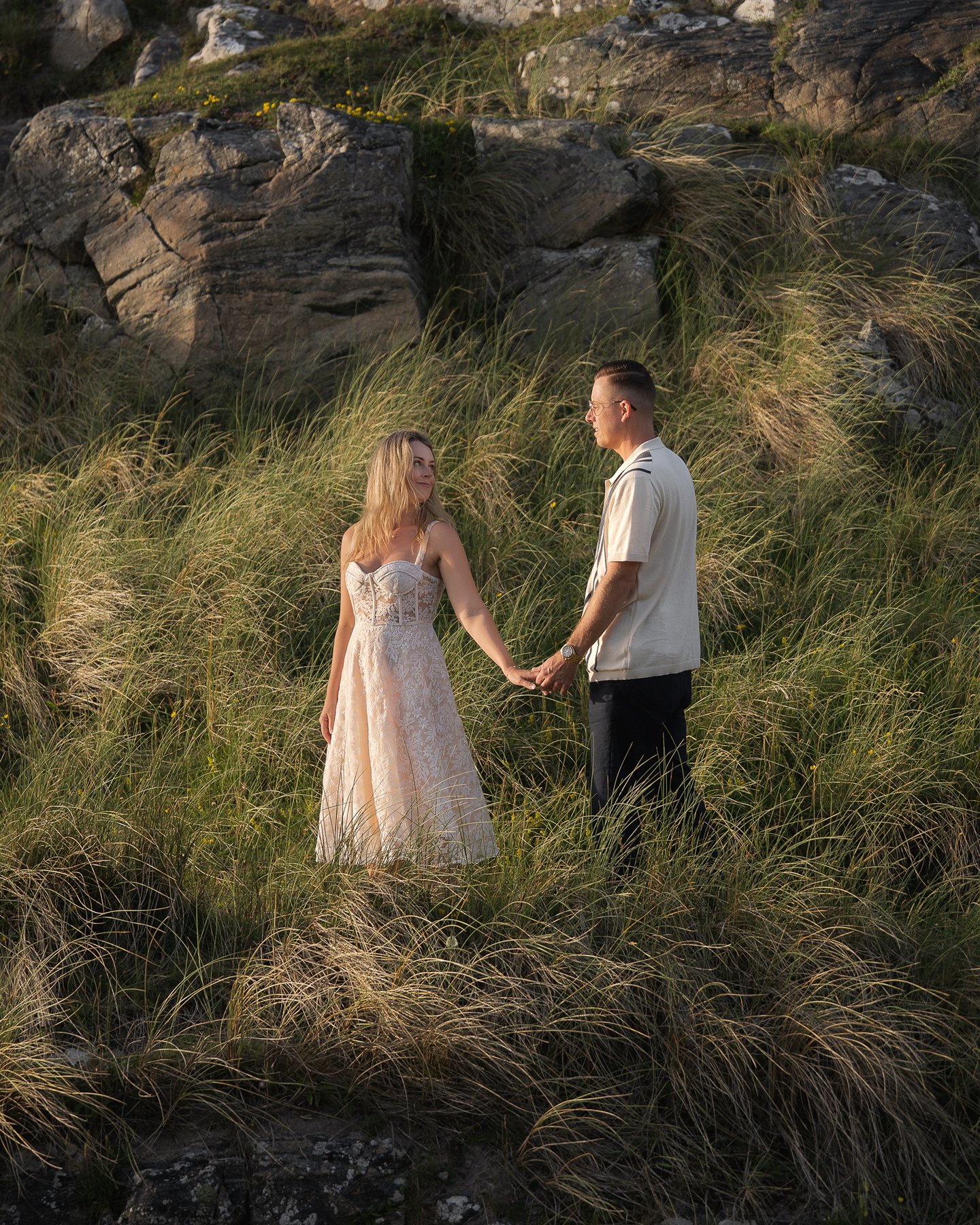 Couple holding hands in tall grass near rocky coastline, woman in white dress during golden hour light.