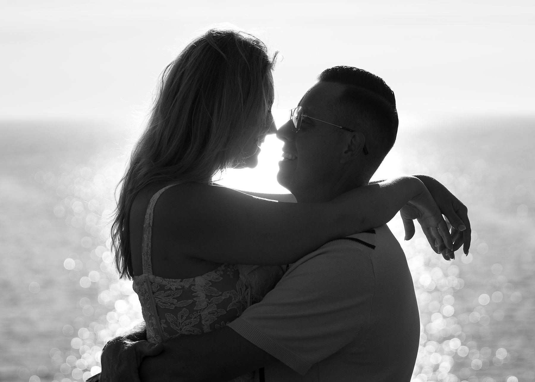 Silhouetted couple holding each other close against sparkling water with sunlight creating dramatic backlighting.