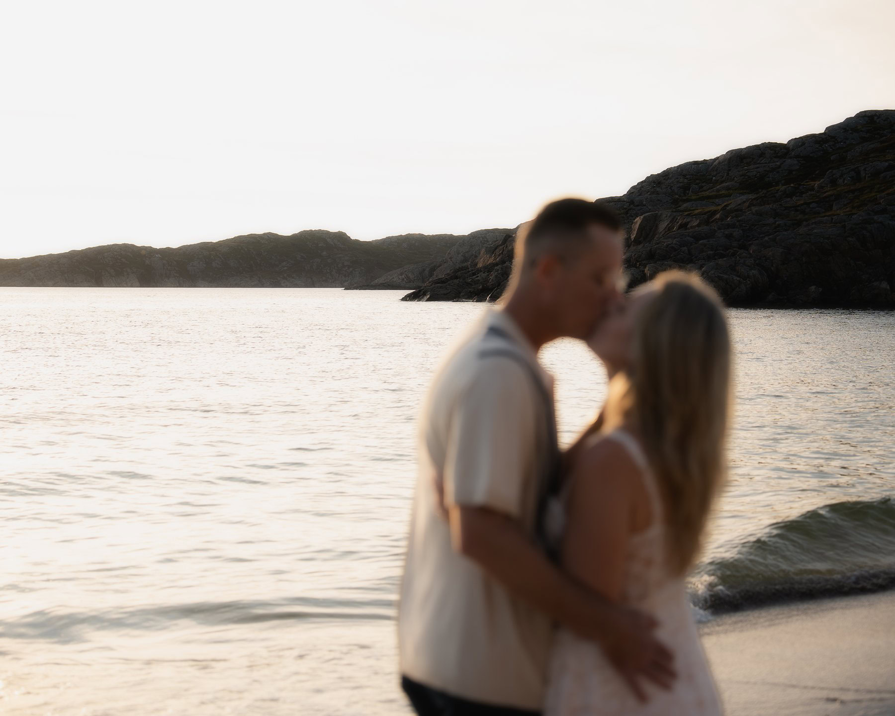 Couple kissing at sunset on a sandy beach in Scotland with waves and rocky cliffs in the background, romantic engagement moment.