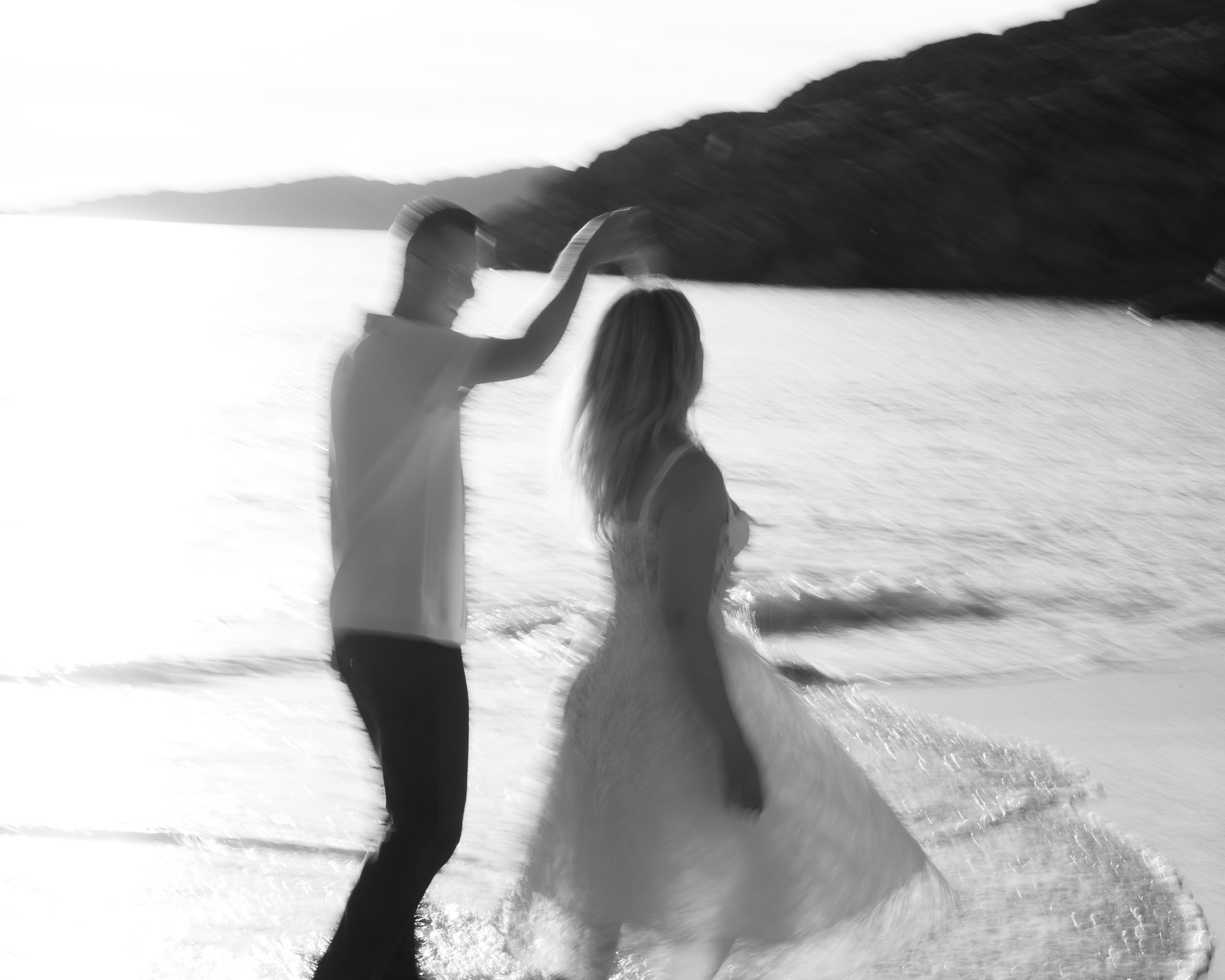 Couple dancing on the beach at sunset with a rocky shoreline in the background, captured in a blurred style.