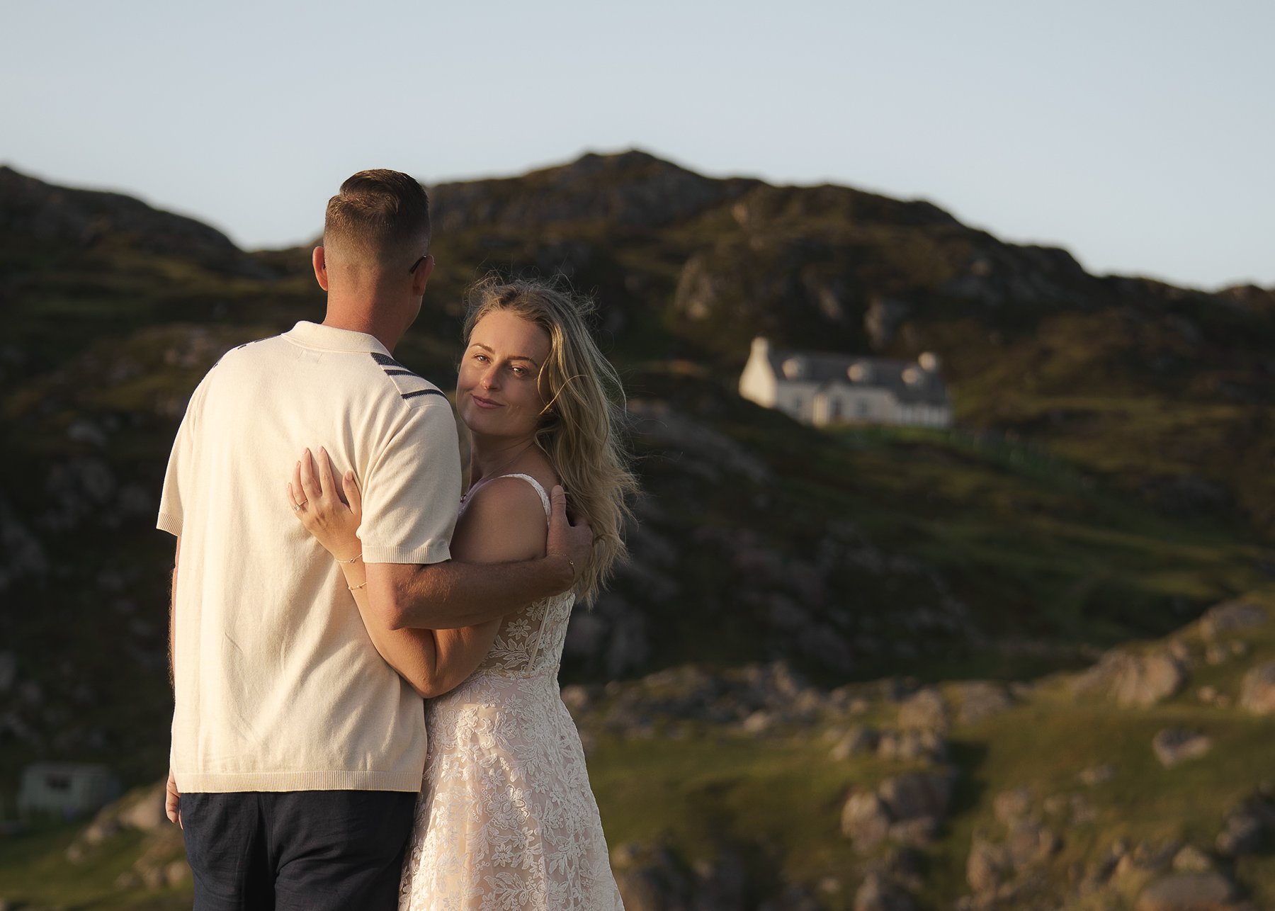 Couple embracing at sunset in Achmelvich Bay, with a white cottage in the hills behind.