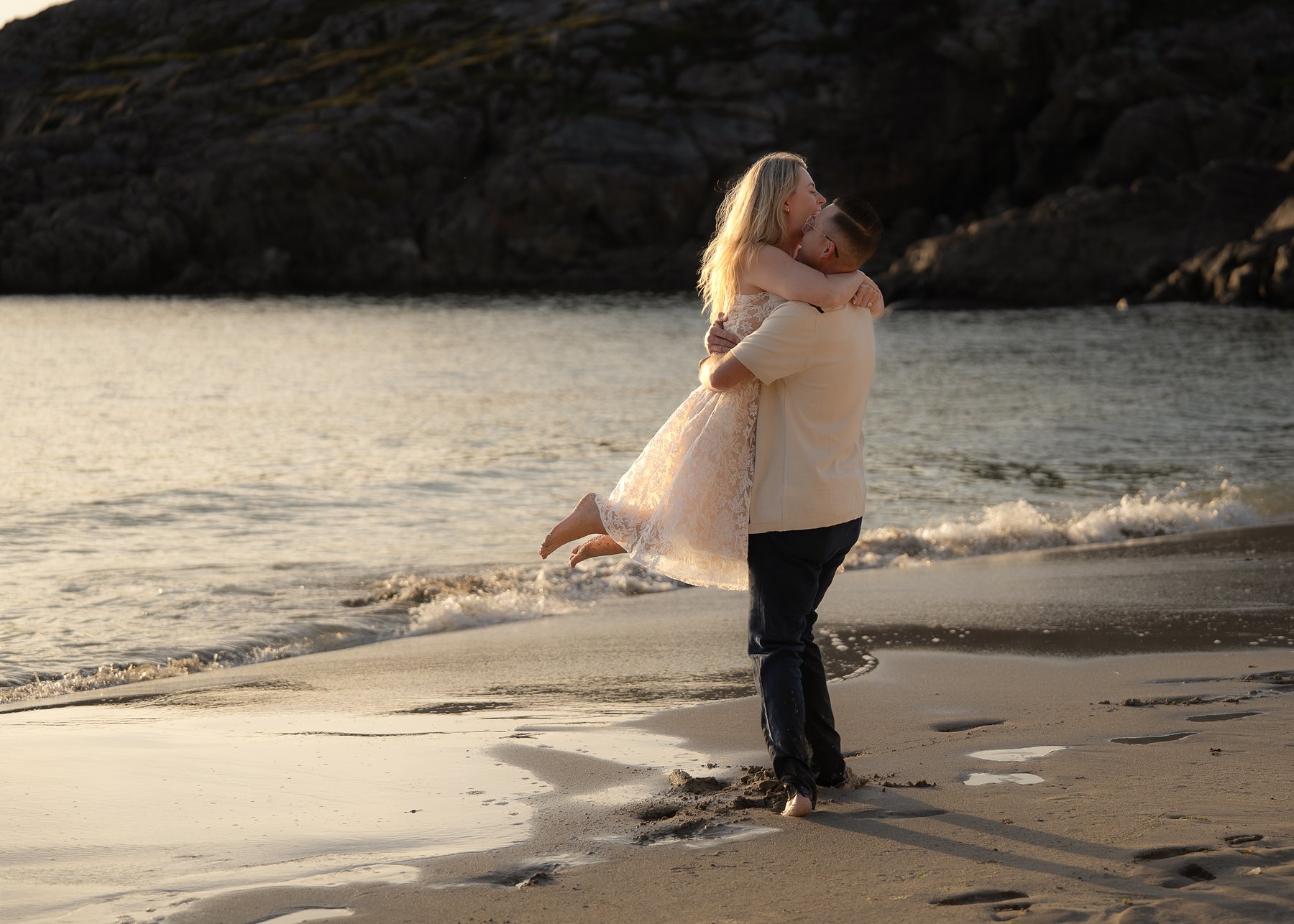 Groom lifting bride on Scottish beach during golden hour engagement photoshoot with waves and cliffs