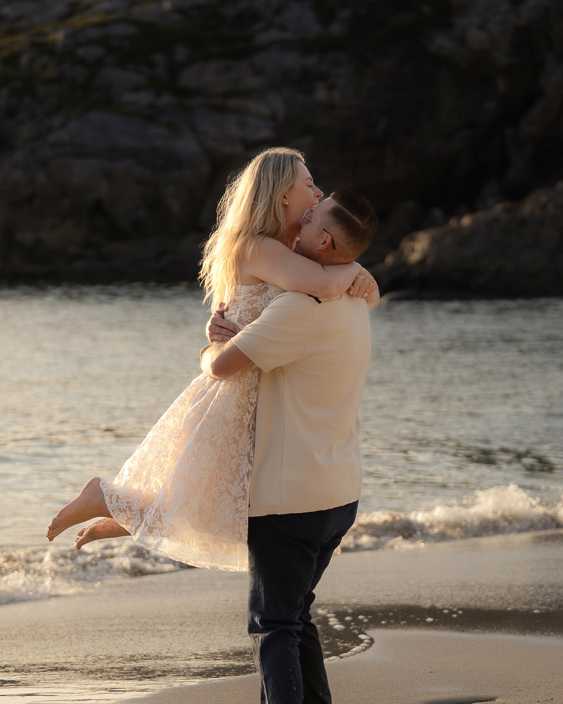Man lifting woman in joyful embrace on sandy beach with waves and dramatic rocky cliffs in background.