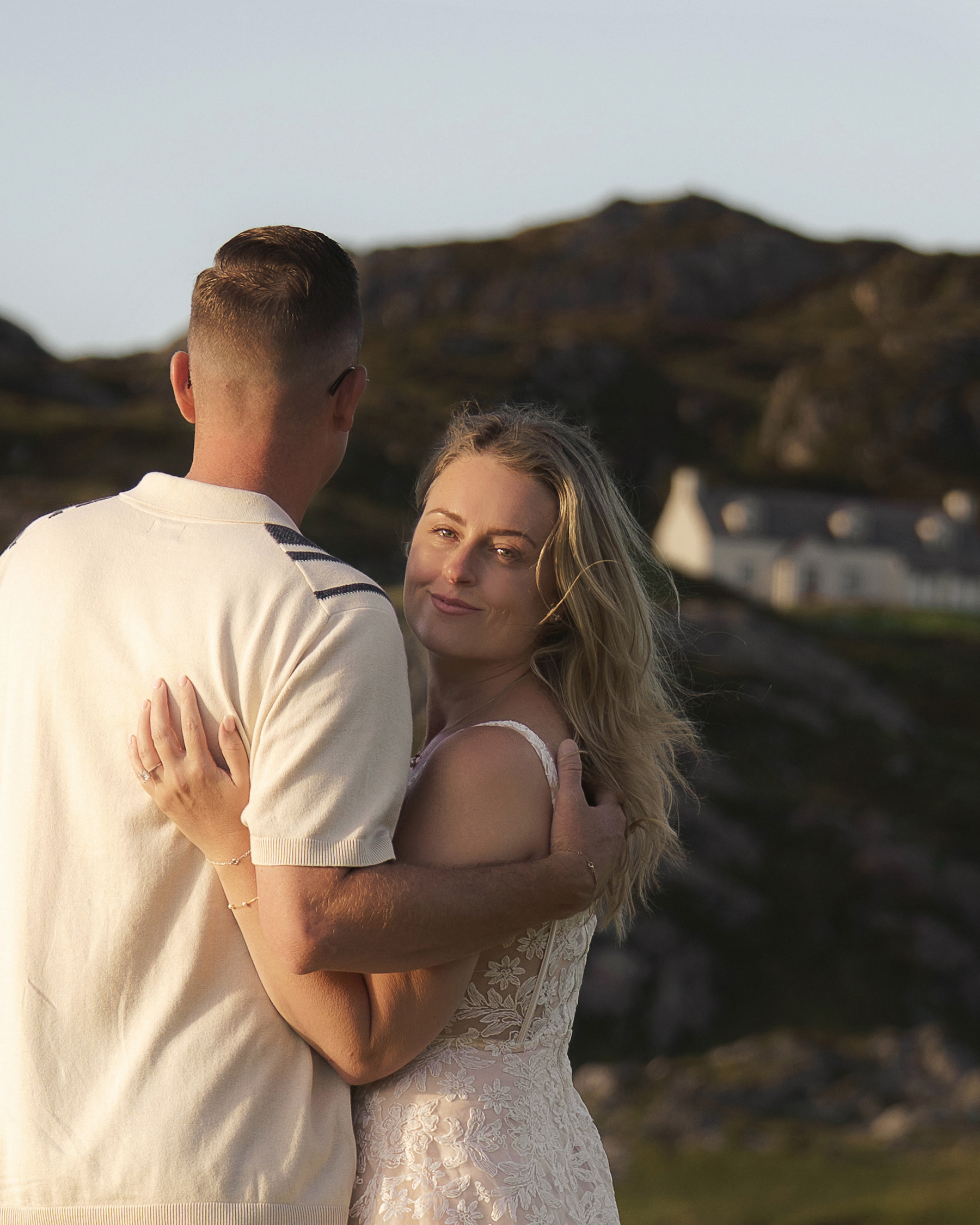 Couple hugging in Highland landscape with blonde woman looking at camera and white cottage in distance.