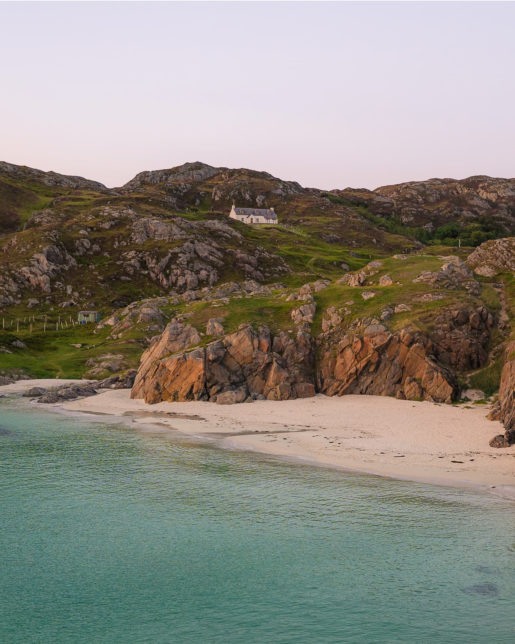 Pristine turquoise bay with white sand beach surrounded by dramatic rocky cliffs and remote cottage on hilltop.