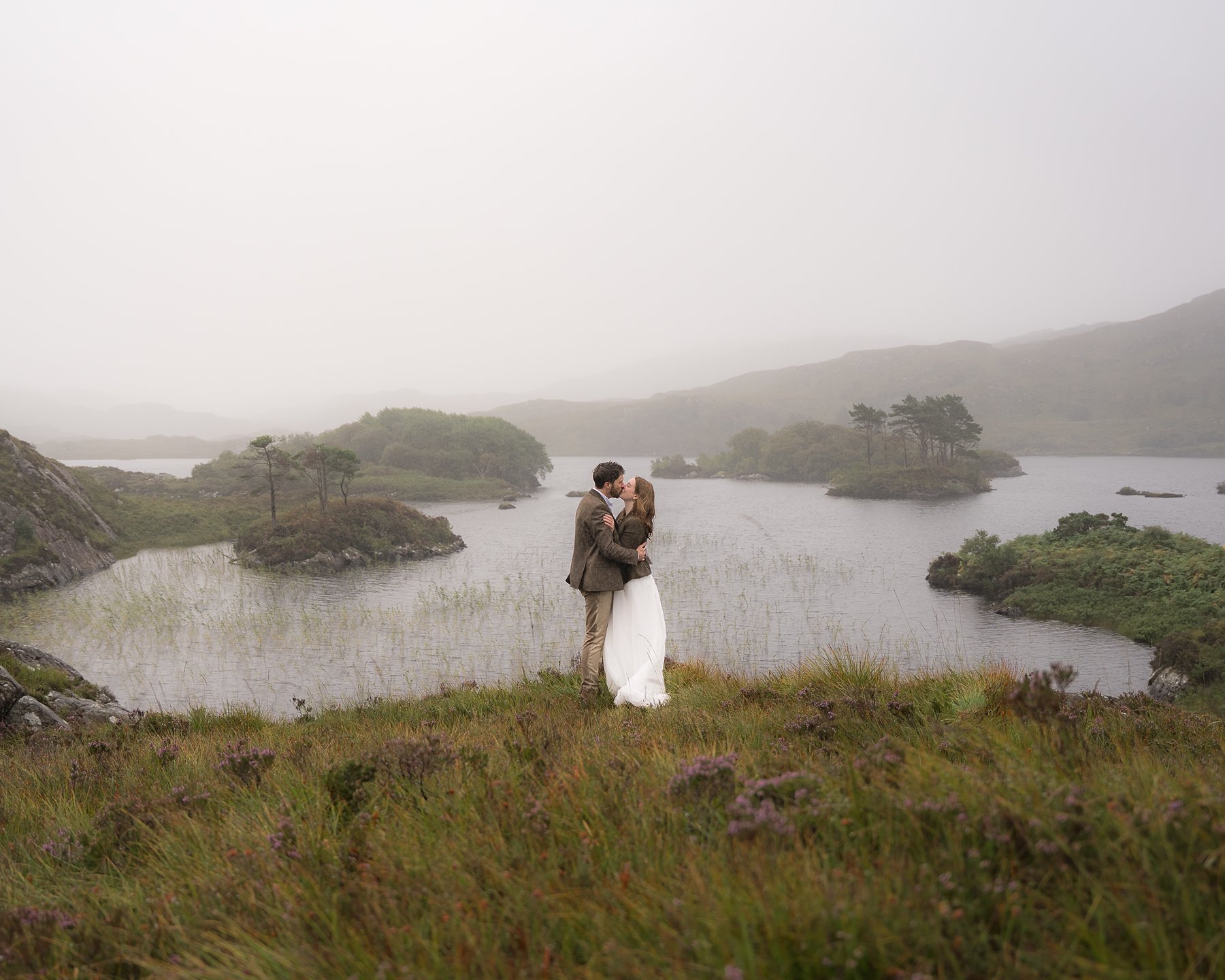 Bride and groom kissing on hillside overlooking misty loch with tree-covered islands.