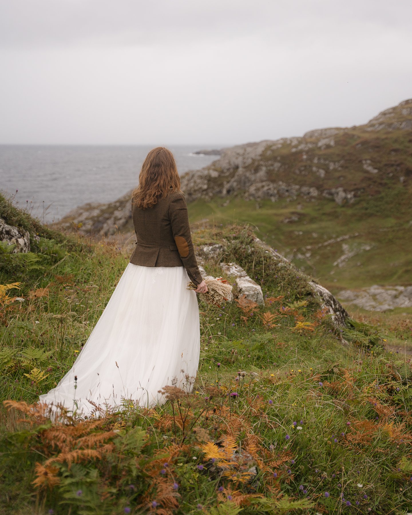 Woman in white dress and brown jacket standing on rugged coastal clifftop overlooking ocean on overcast day.