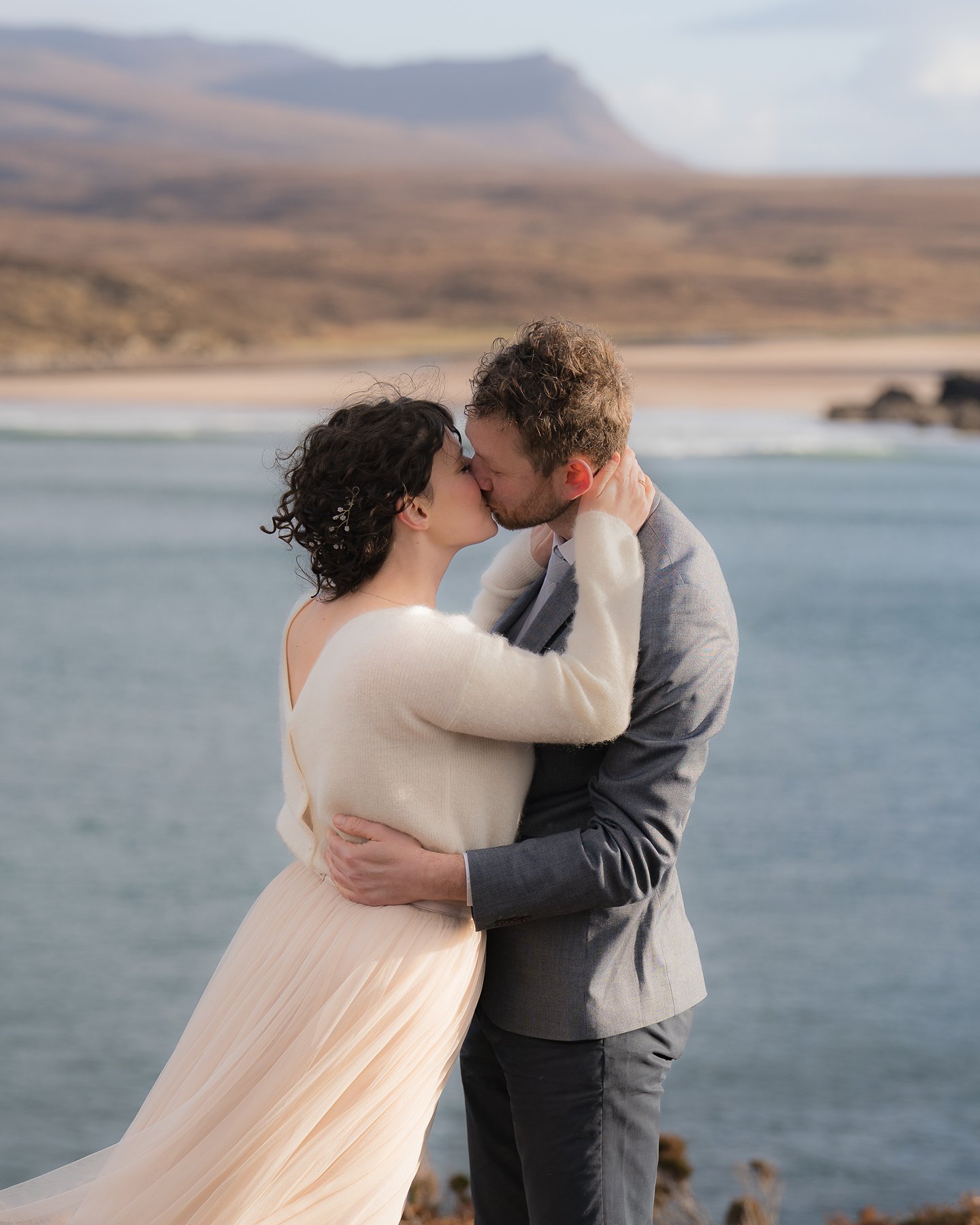 Elopement couple  kissing and embracing by sea with beach and mountains in background.