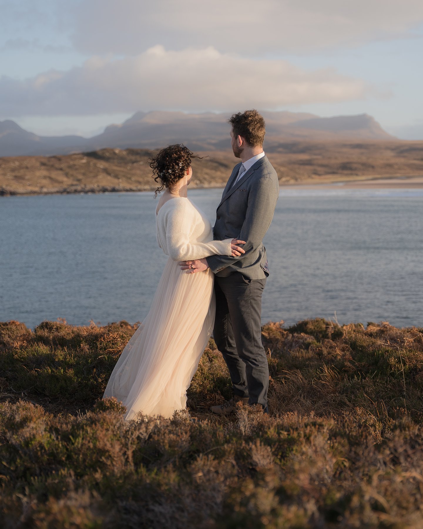Bride and groom standing by a sea, gazing at Scottish Highlands mountains on their intimate wedding day.