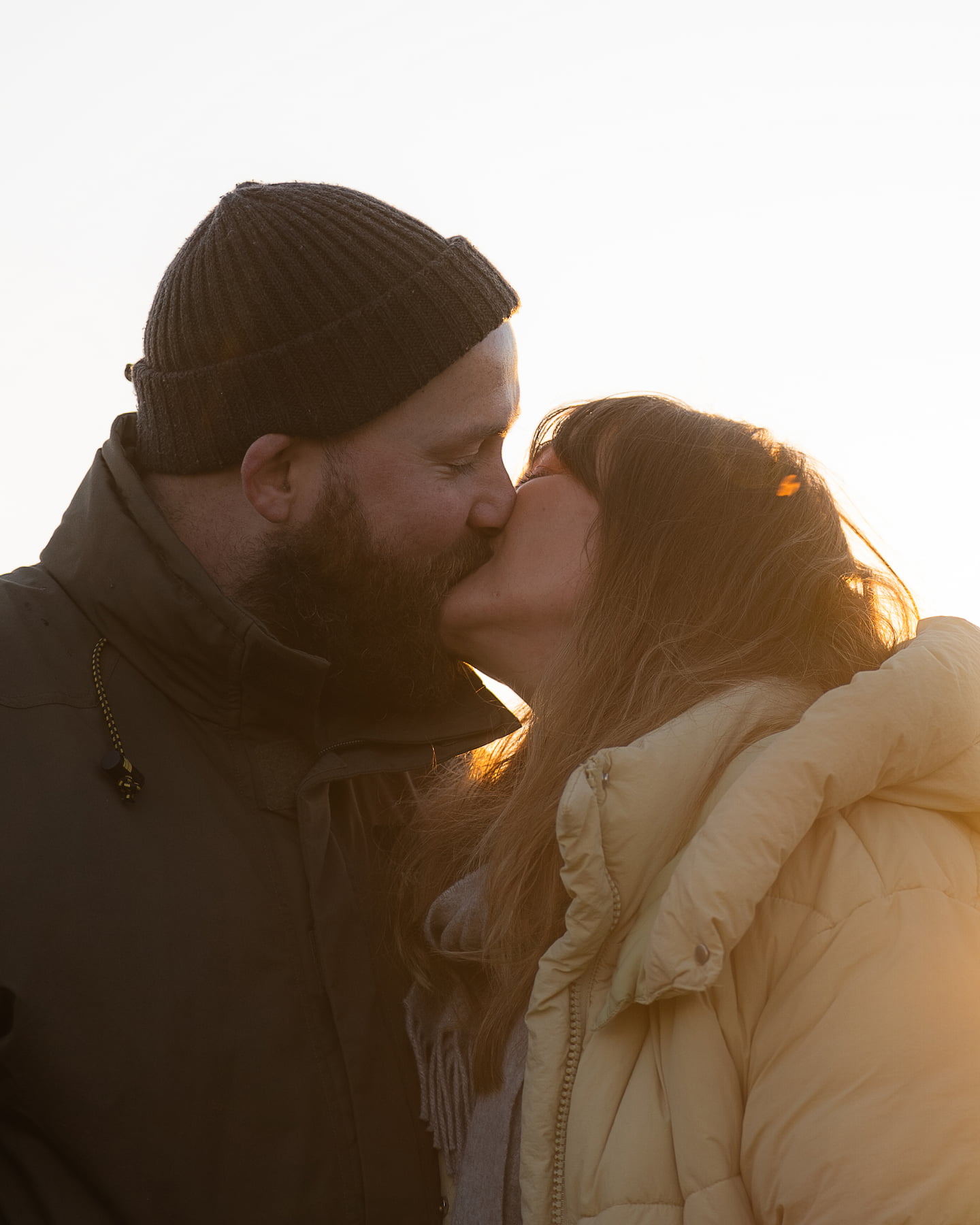 Couple kissing at sunset during outdoor photoshoot, wearing winter jackets.